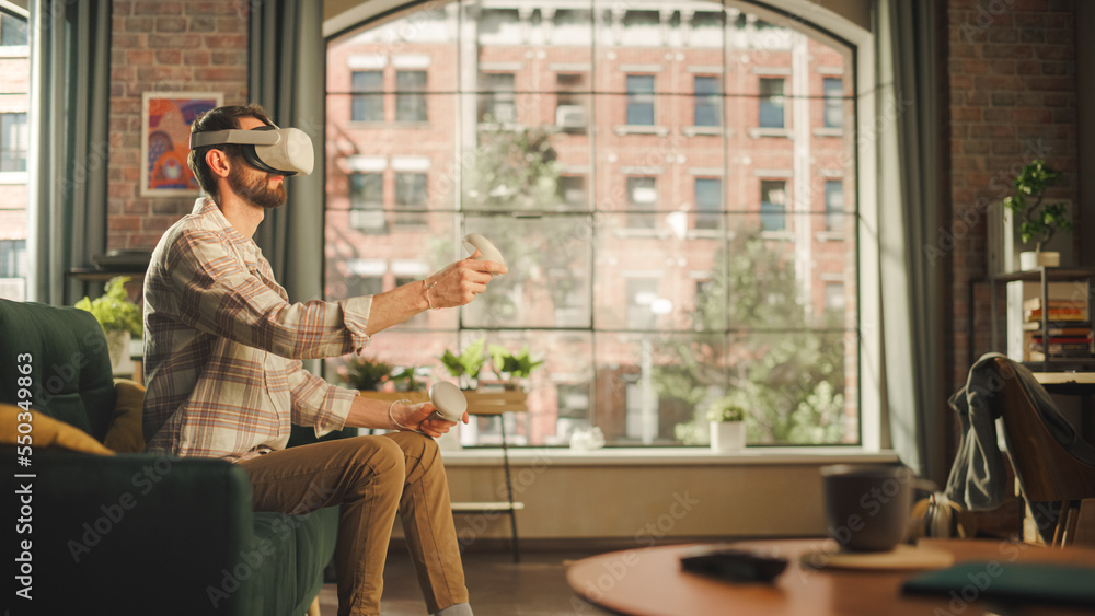 Adult Bearded Male Using Virtual Reality Headset and Controllers at Home. Creative Man Sitting on a 