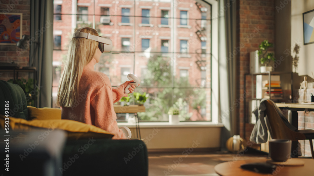 Female with Blond Hair Using Virtual Reality Headset and Controllers at Home. Creative Woman Sitting