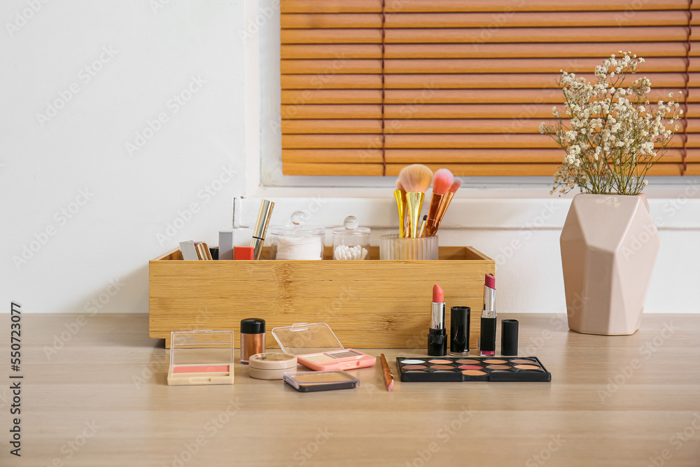 Box with makeup brushes, decorative cosmetics and flowers in vase on table near light wall