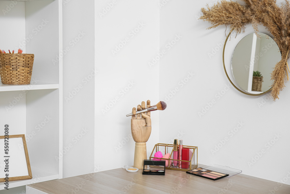 Holders with makeup brush and decorative cosmetics on table near light wall