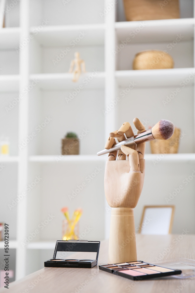 Wooden hand with makeup brush and eyeshadows on table in room, closeup