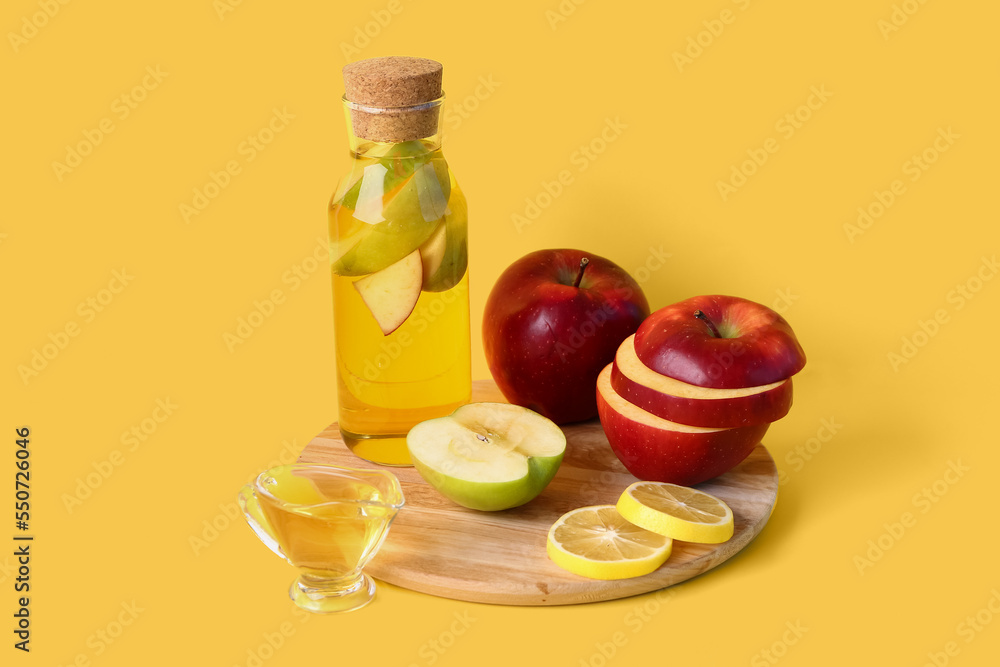 Wooden board with bottle of apple cider vinegar and fresh fruits on yellow background