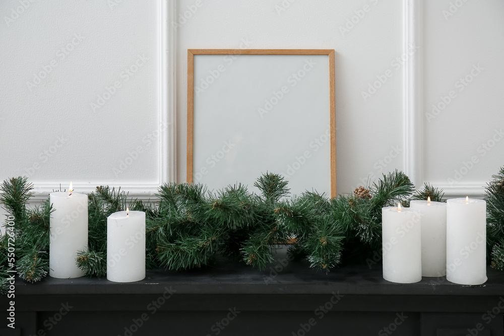 Blank frame with Christmas branches and candles on mantelpiece near light wall