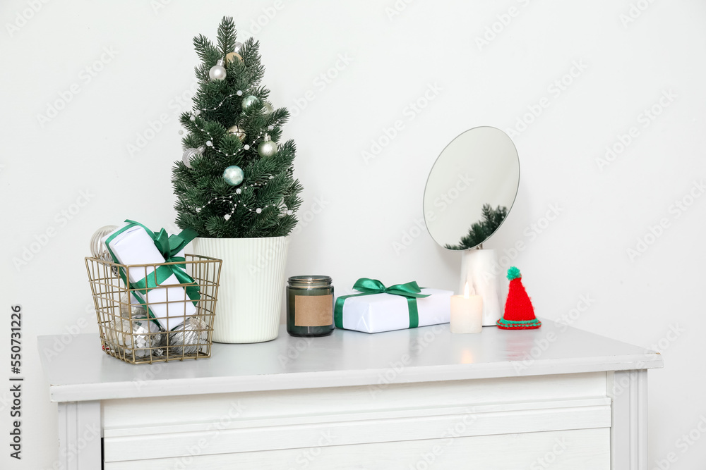 Small Christmas tree with presents, candles and mirror on shelf near light wall