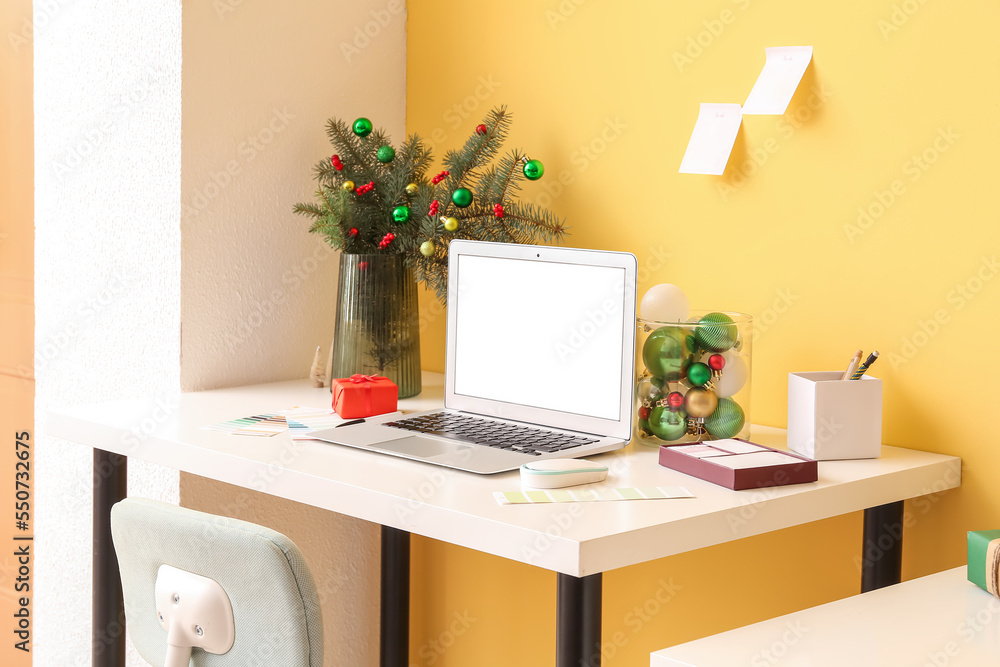 Workplace with laptop, fir branches in vase and Christmas presents near yellow wall