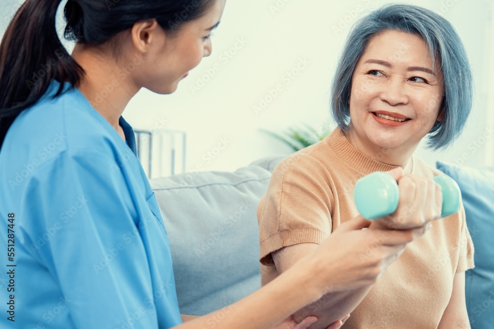 Caregiver helping contented senior woman exercise with dumbbell at home. Rehab for elder, senior med