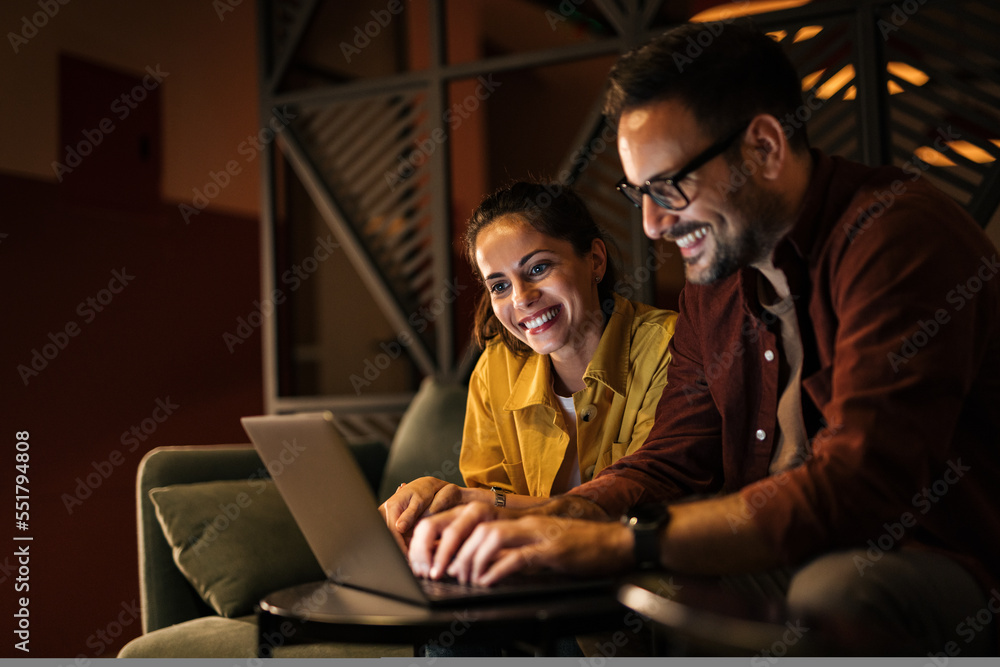 Happy man and woman working overtime, using a laptop, sitting at the office.