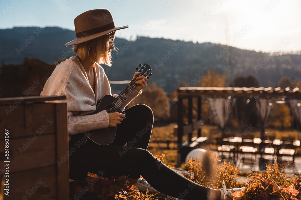 Girl in cowboy hat playing ukulele sitting in home garden patio