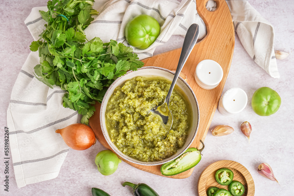 Wooden board with bowl of tasty green salsa sauce and ingredients on light background