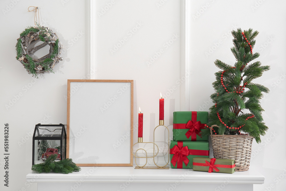 Blank frame with Christmas tree, candles and presents on mantelpiece near light wall