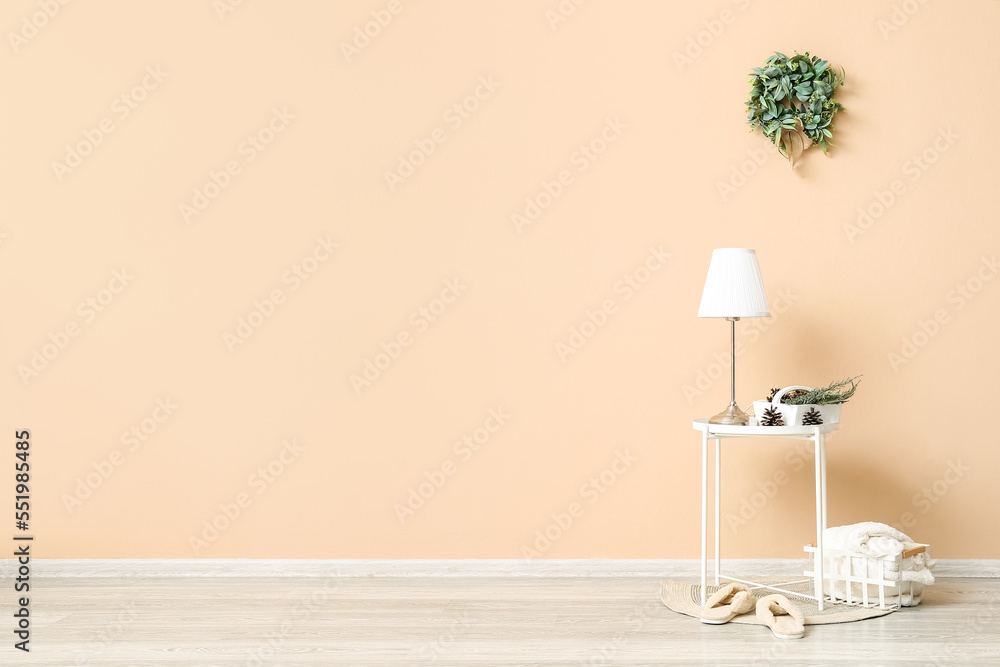 Table with lamp, slippers and Christmas mistletoe wreath on beige wall in room