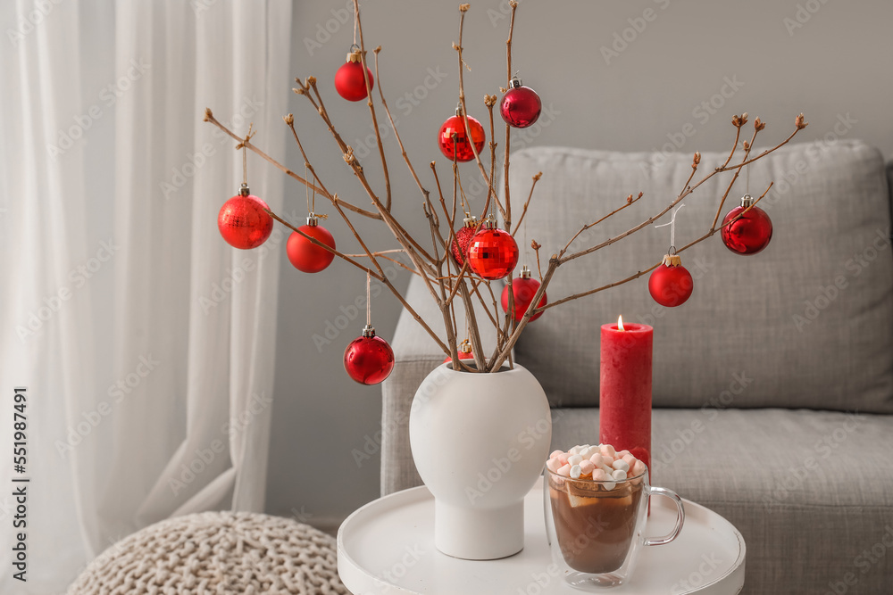 Vase with tree branches, Christmas balls, candle and cup of cocoa on table in living room