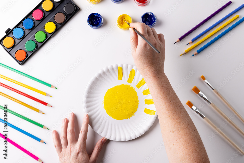 Overhead of hands painting with yellow paint on paper plate, with art materials on table top