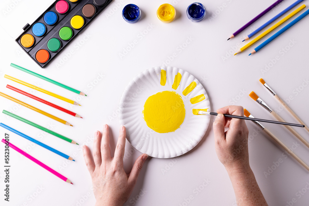 Overhead of hands painting with yellow paint on paper plate, with art materials on table top