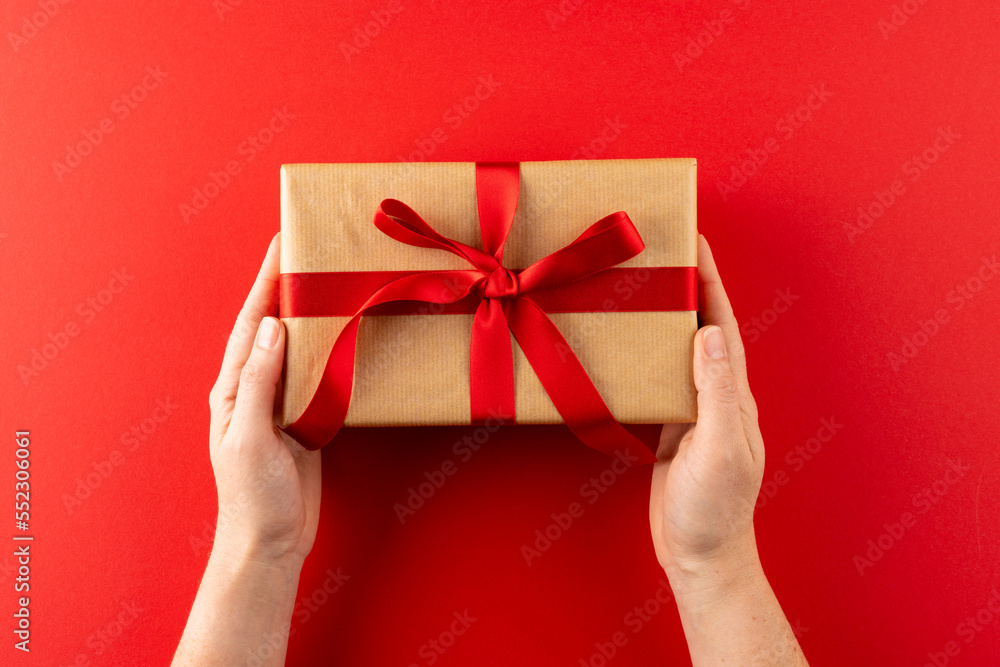 Overhead of hands holding gift in brown paper with red ribbon, on red background with copy space