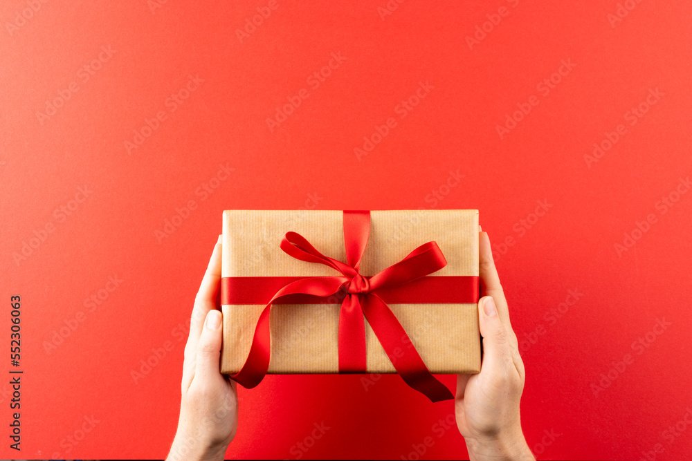 Overhead of hands holding gift in brown paper with red ribbon, on red background with copy space