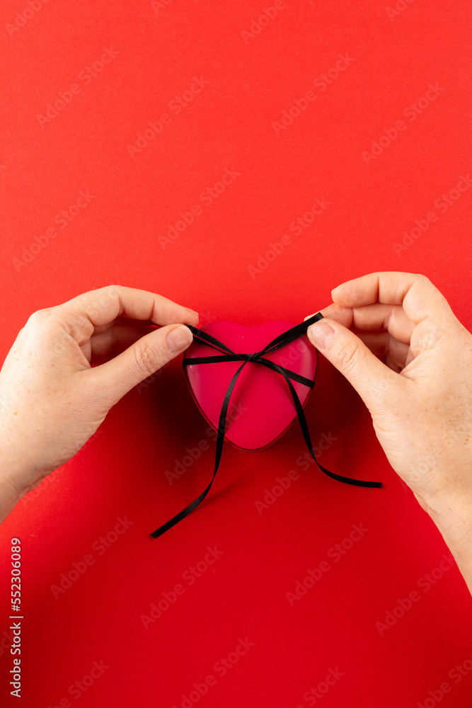 Vertical of hands tying red heart gift box with black ribbon, on red background with copy space