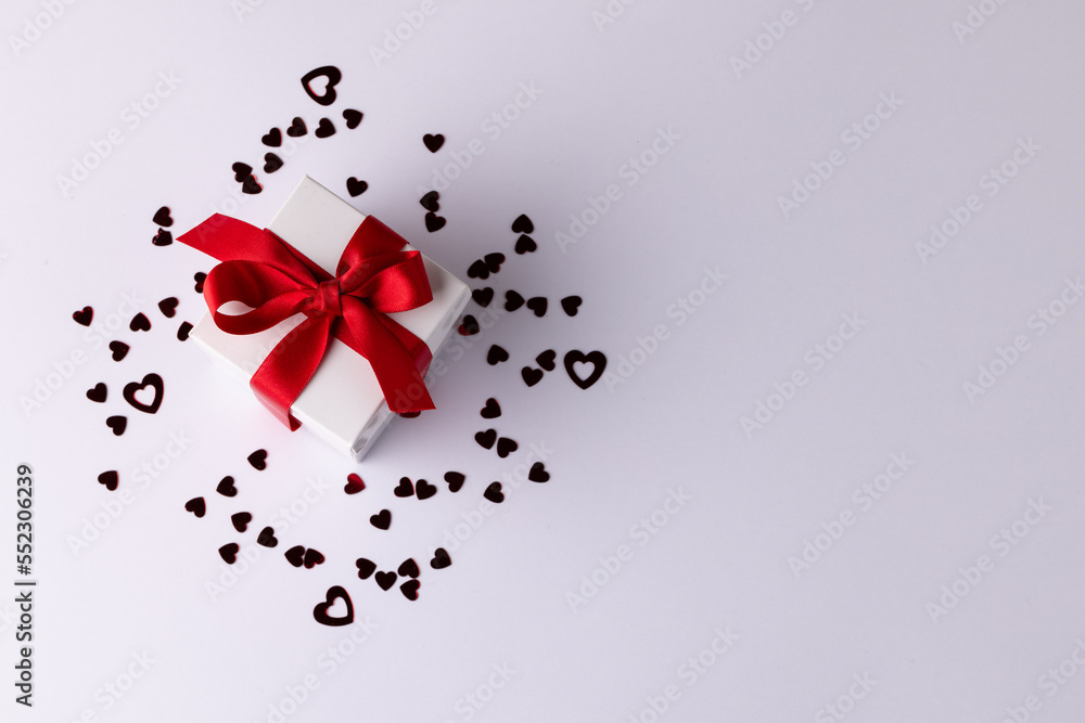 White gift box with red ribbon on heart confetti, on white background with copy space