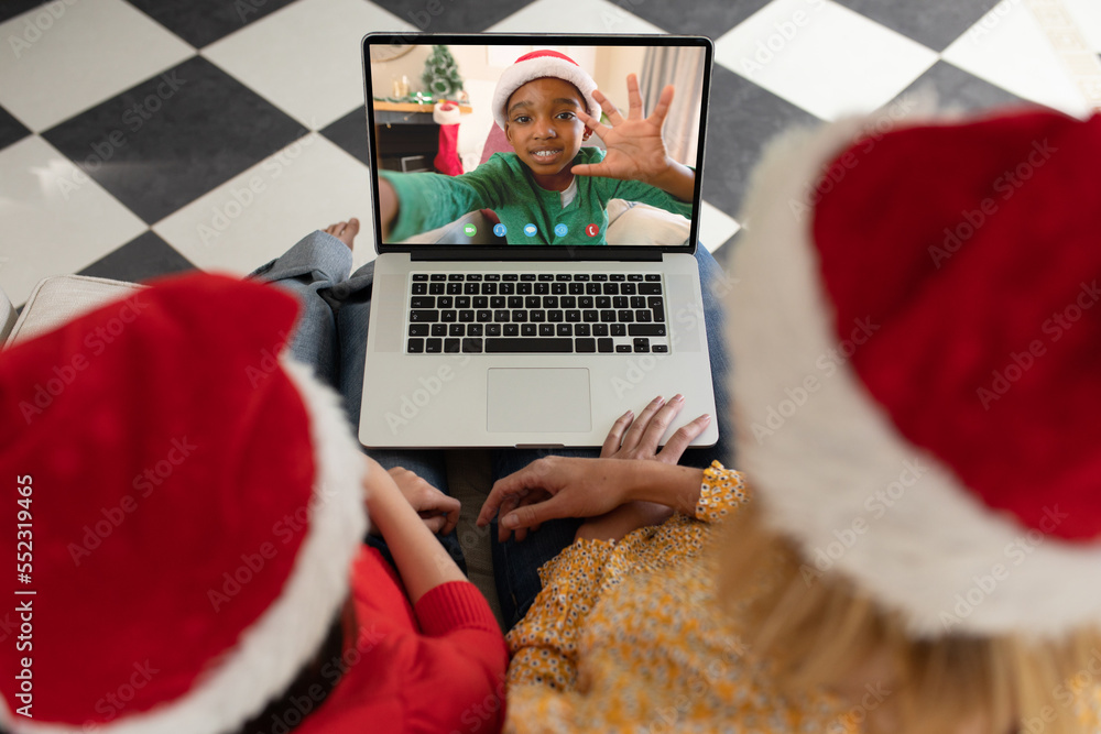 Diverse mother and daughter with santa hats having video call with happy african american boy