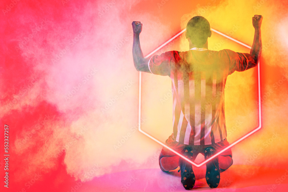 African american male soccer player with arms raised kneeling by illuminated hexagon