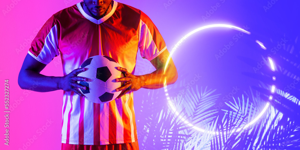 African american male soccer player holding ball by illuminated circle and plants, copy space