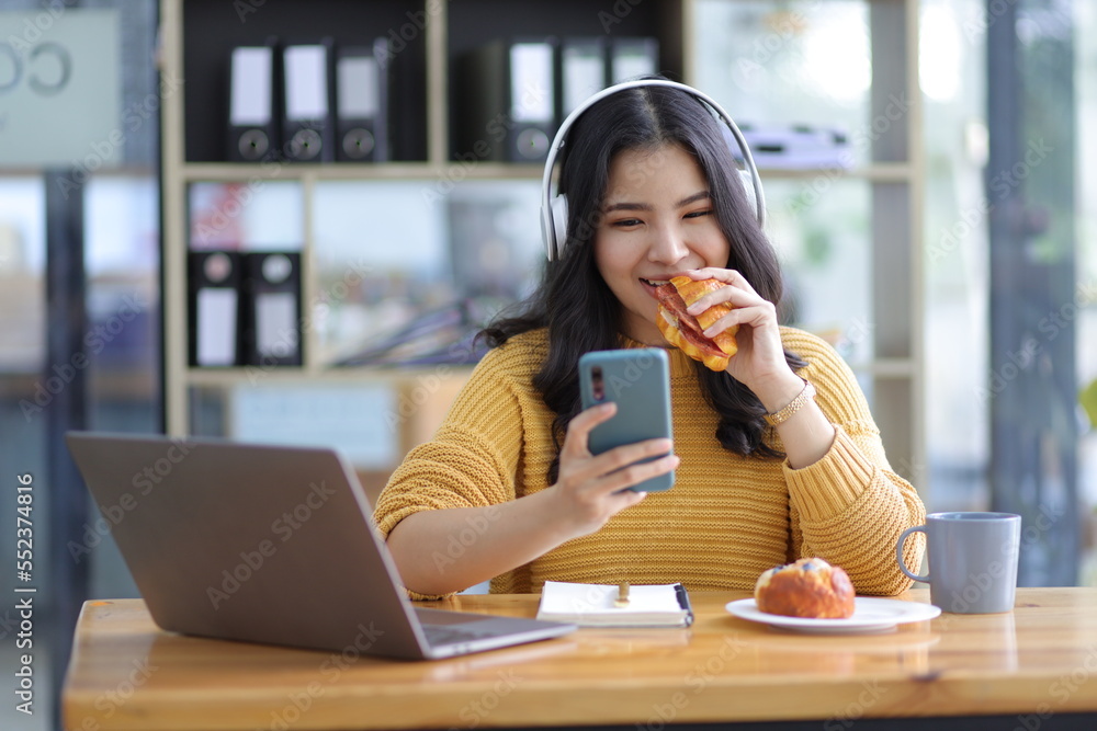 迷人的亚洲美女，在办公室里随意地用智能手机工作，吃零食。