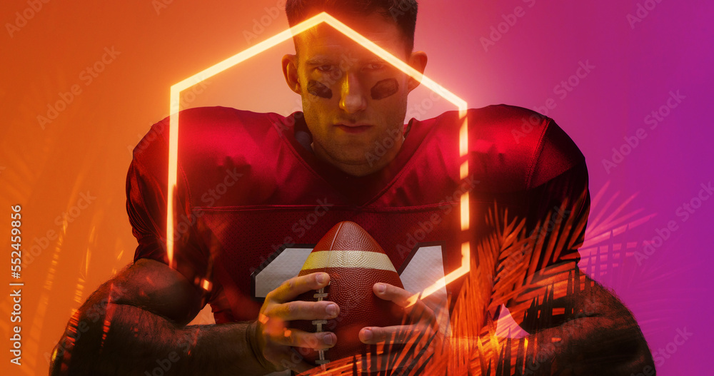 Portrait of american football player with black paint on face holding ball by hexagon and plants