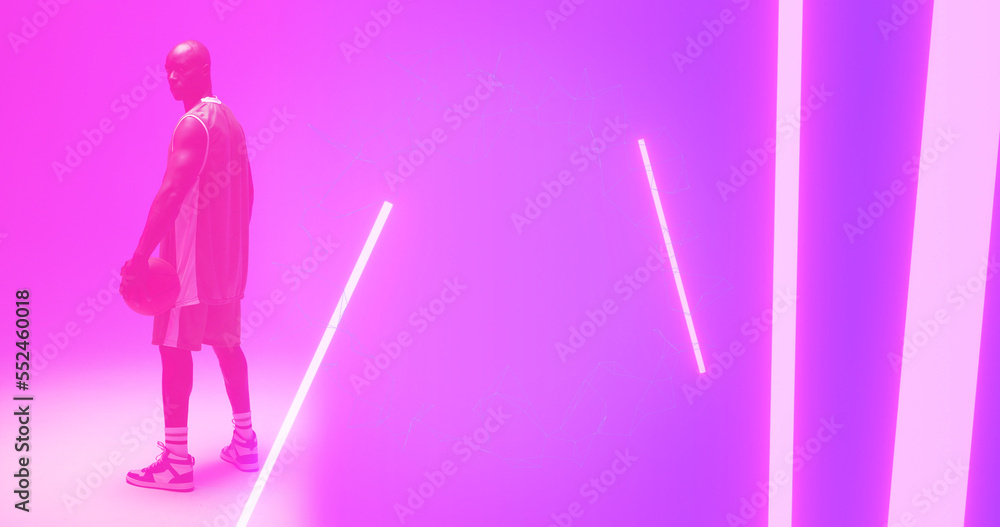 Bald african american basketball player with ball standing by illuminated lights on pink background