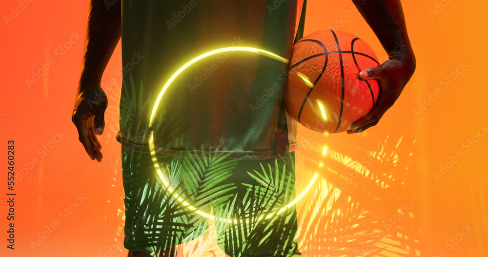 Midsection of african american basketball player holding ball by illuminated circle and plants