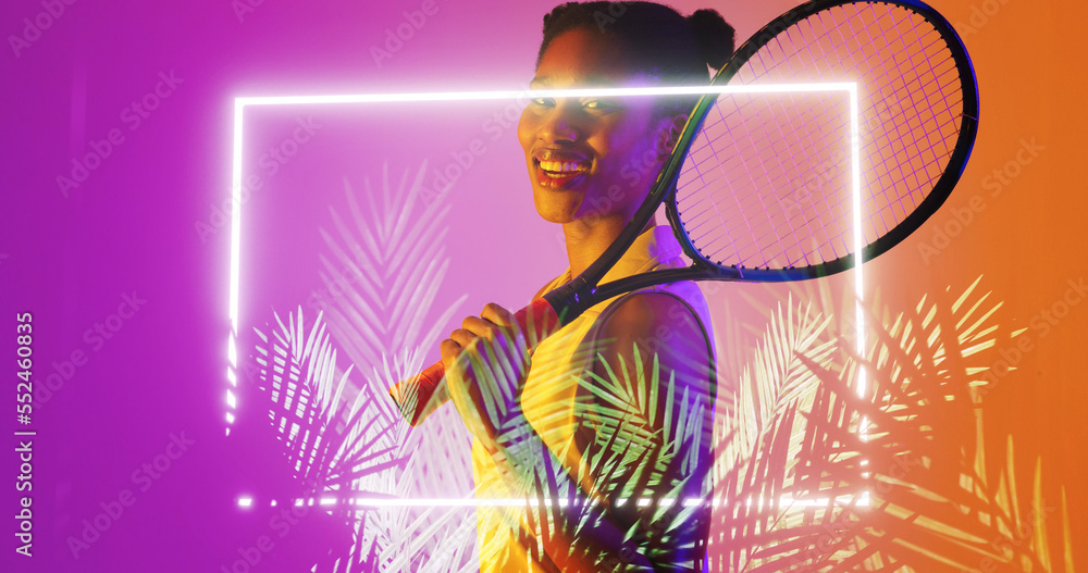 Biracial smiling female player with tennis racket standing by illuminated rectangle and plants