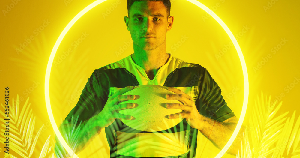 Caucasian male player holding rugby ball over illuminated circle and plants on yellow background