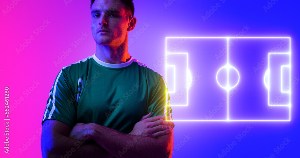 Confident caucasian player with arms crossed by neon soccer field over blue and pink background