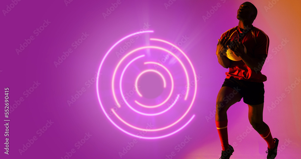 African american rugby player holding ball by illuminated circular pattern on purple background