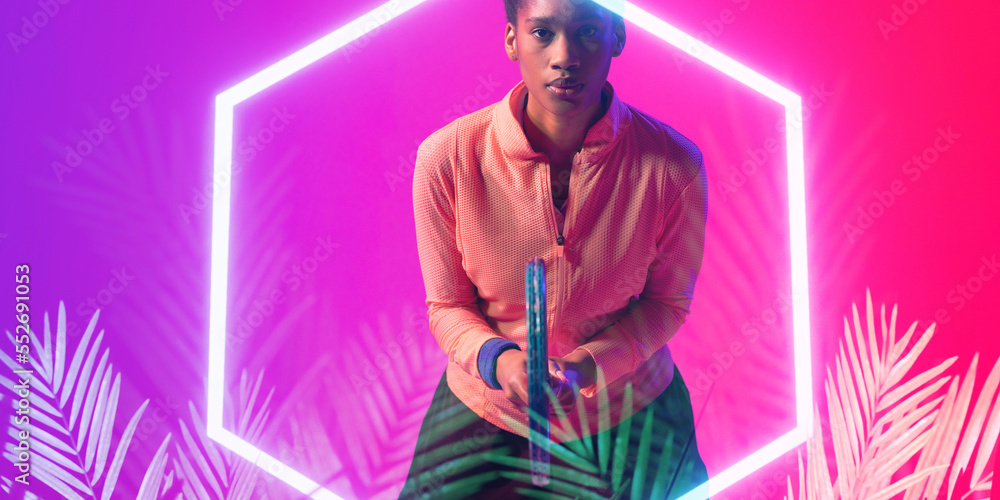 African american female tennis player in hitting stance holding racket by glowing hexagon and plants