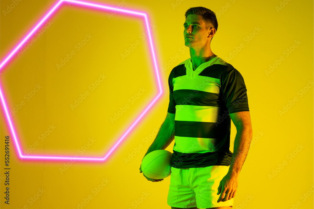 Caucasian male rugby player with ball standing by illuminated hexagon over yellow background