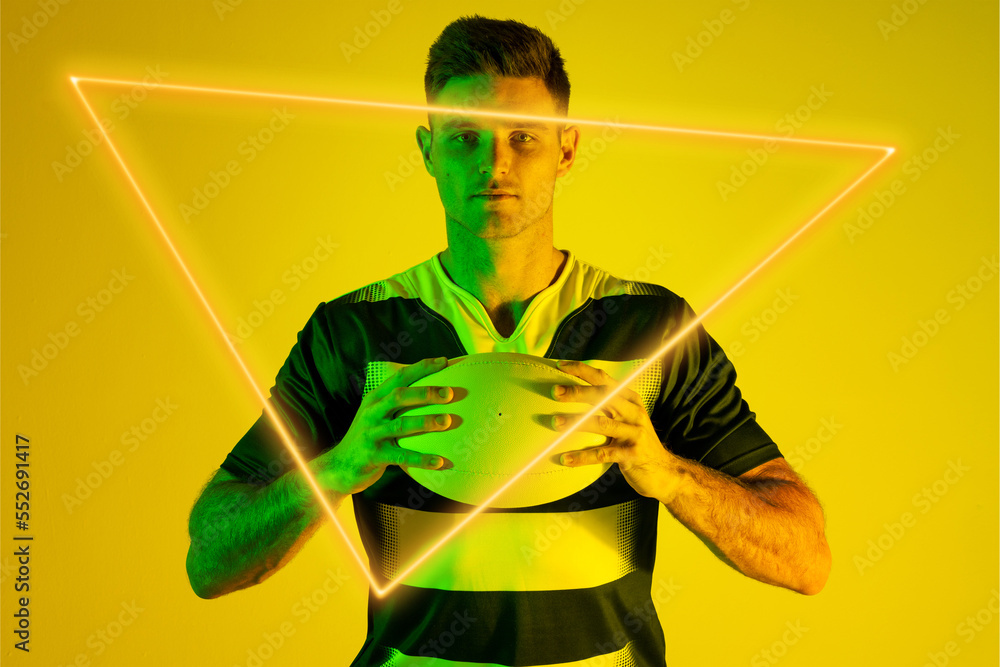 Portrait of caucasian male rugby player holding ball by illuminated triangle on yellow background