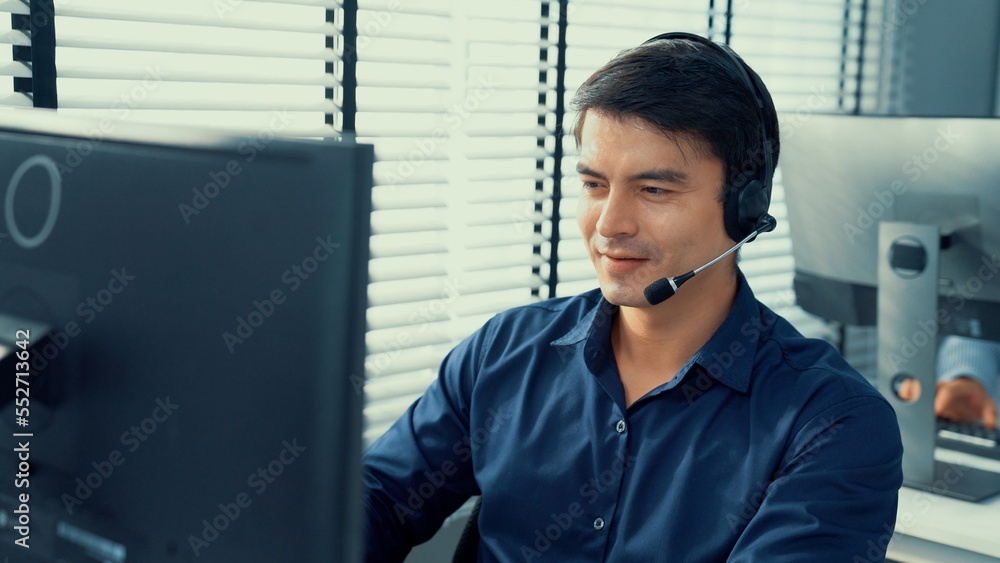 Young competent asian male call center agent working at his computer while simultaneously speaking w