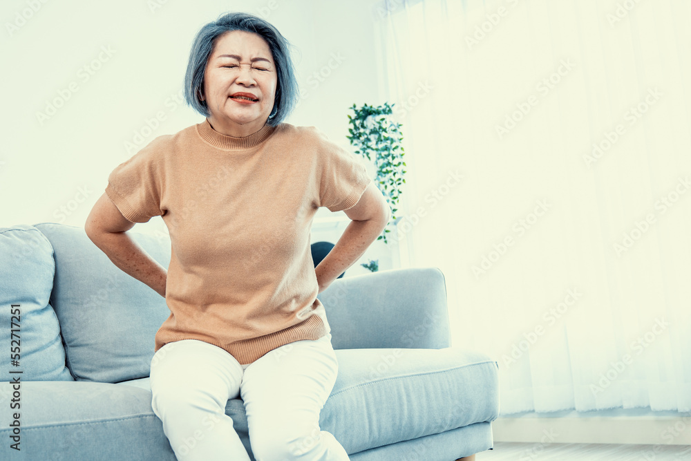 An agonizing senior woman experiencing back pain while sitting on her sofa at home and holding her b