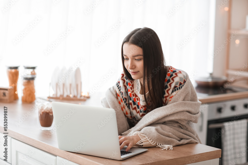 Young woman in warm plaid using laptop in kitchen