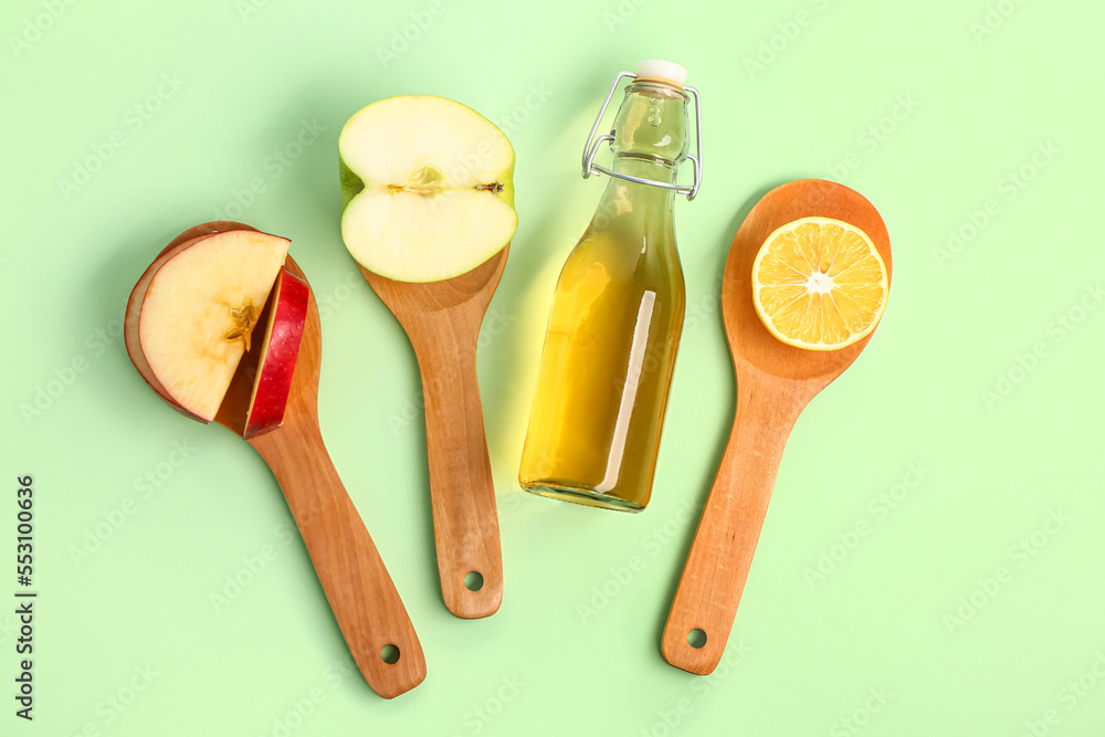 Bottle of apple cider vinegar and spoons with fruits on color background