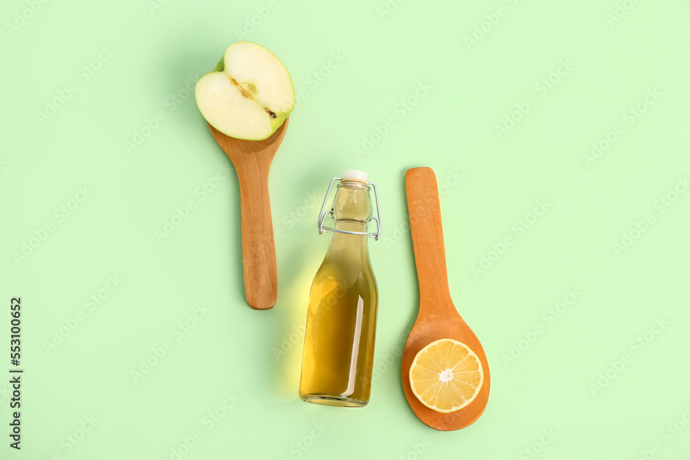 Bottle of apple cider vinegar and spoons with fruits on color background