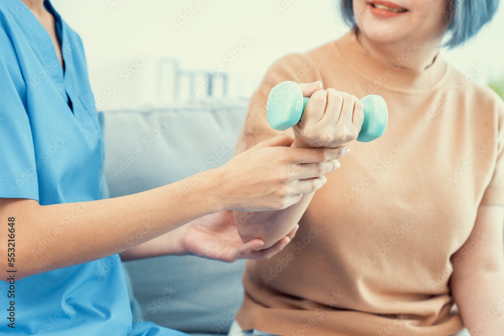 Caregiver helping contented senior woman exercise with dumbbell at home. Rehab for elder, senior med