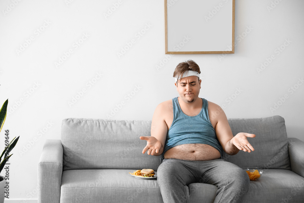 Upset young overweight man with unhealthy food sitting on sofa at home