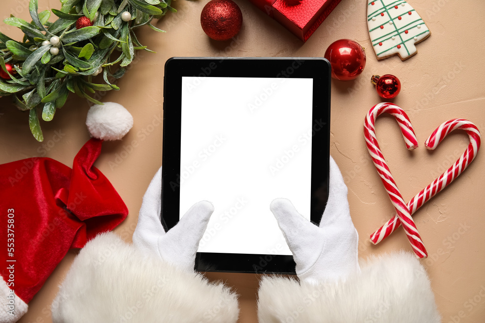 Santa Claus with tablet computer, hat and Christmas decor on beige background