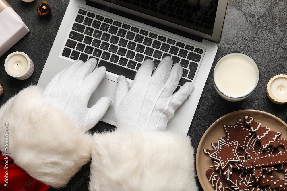 Santa Claus with glass of milk and cookies using laptop on black background