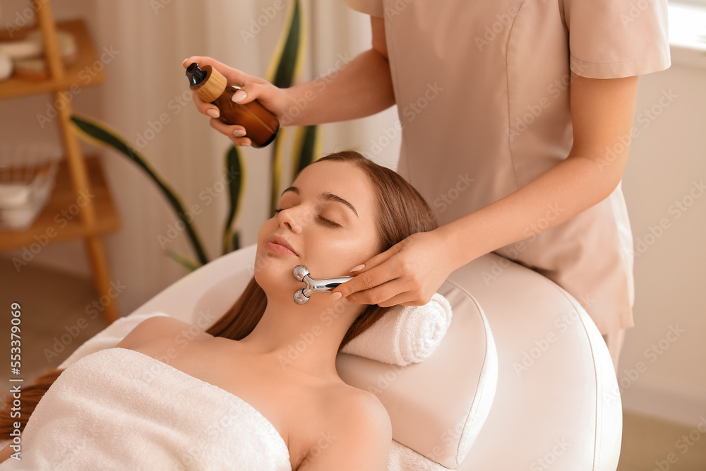 Pretty young woman receiving face massage with roller in beauty salon