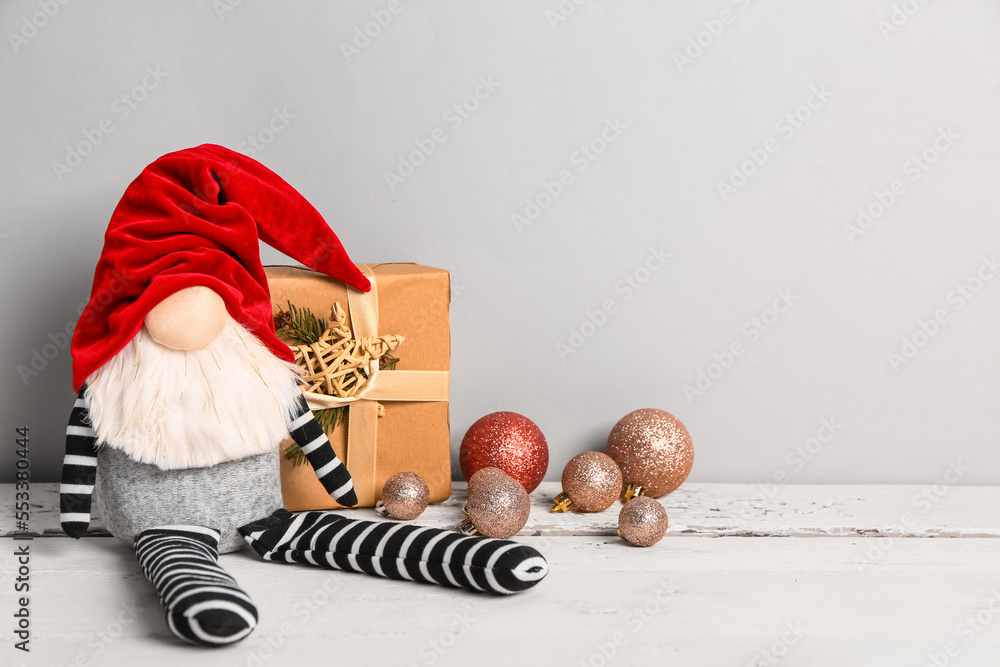 Christmas gnome, gift box and balls on light wooden table against grey background