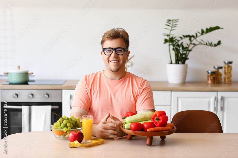 Young overweight man with healthy food, glass of juice and measuring tape at table in kitchen