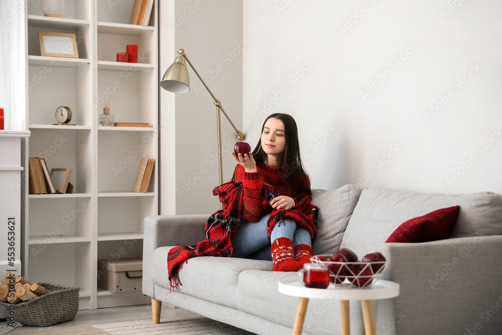 Young woman in warm sweater with apple sitting on sofa at home