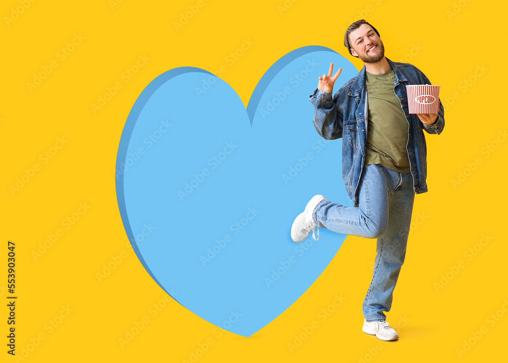 Happy young guy with bucket of popcorn and big heart on yellow background